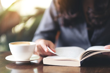 Closeup image of a woman holding and reading a book with coffee cup on wooden table