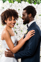 cheerful african american bride hugging happy bridegroom near flowers
