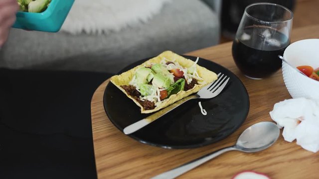 Putting some avocado on vegan minced soy meat laying on a hard shell tortilla. Showing extra ingredients in the background.