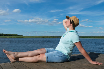 Fototapeta na wymiar girl in a hat sunbathes on a pier by the lake on a hot sunny day