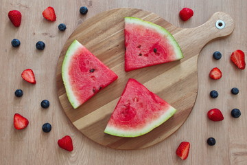 Directly above shot of watermelon slices, strawberries and blueberries on the cutting board against the wooden background
