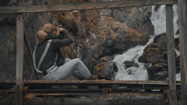 Girl tourist sitting on a wooden bridge with a camera in his hands and taking pictures of the waterfall in the rocks. Free travel, girl photographer takes a picture of the landscape. Slow motion, 4K