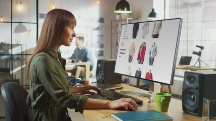 Beautiful Brunette Female Scrolls Through Online Fashion Store on Her Personal Computer with Big Display. She Works in a Cool Office Loft. Other Male Creative Colleague Walks in the Background.
