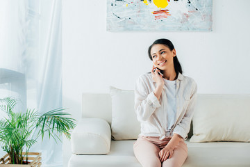 beautiful latin woman talking on smartphone while sitting on sofa and looking away