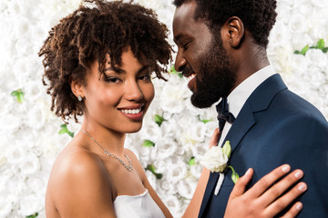 cheerful african american bride hugging handsome bridegroom near flowers - Powered by Adobe