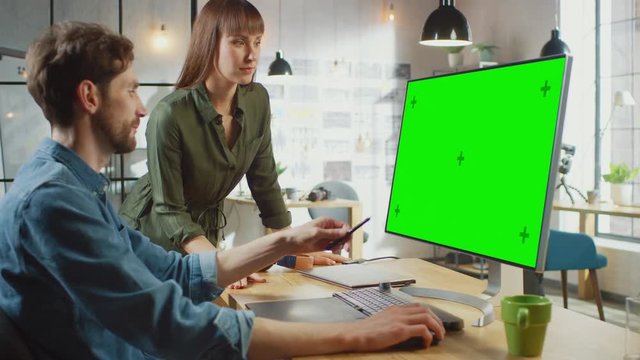Female Art Director Consults Designer Colleague, They Work on a Personal Computer with Green Screen Mock Up Display. They Work in a Cool Office Loft. They Look Very Creative and Cool.