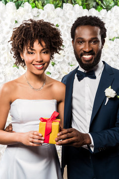 happy african american bride holding present near bridegroom and flowers