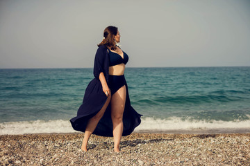 Attractive busty curvy woman in a blue swimsuit resting on the beach. Stylish accessories, fringe, fashion for plus size, beautuful sea. Bodypositive, natural authentic beauty, resort, summer vacation