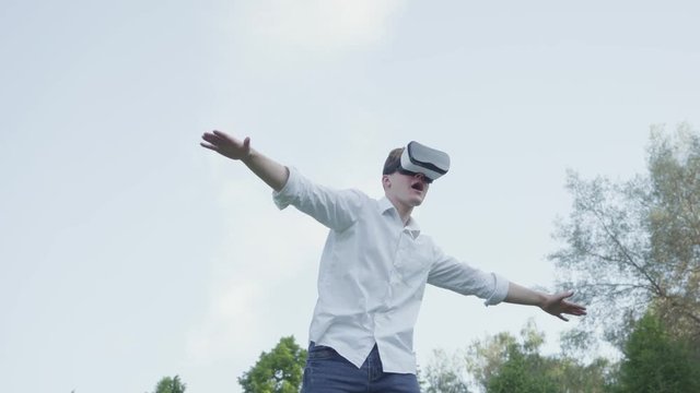 Man wearing virtual reality headset spread his arms to the side, imitating the flight of an airplane in the park enjoying the realistic image. Futuristic and modern technologies. Guy playing game