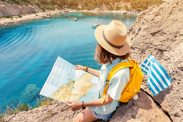Girl tourist student with map and greek flag standing on top of the hill at background of the inspirational blue lagoon bay. Solo Travel in Greece concept