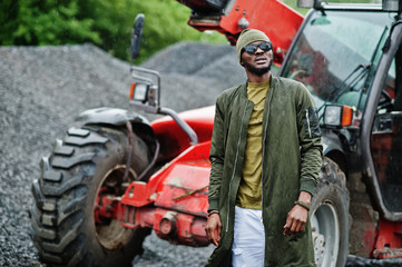 Fototapeta na wymiar Stylish african american man in hat and sunglasses posed outdoor in rain against tractor with a bucket.