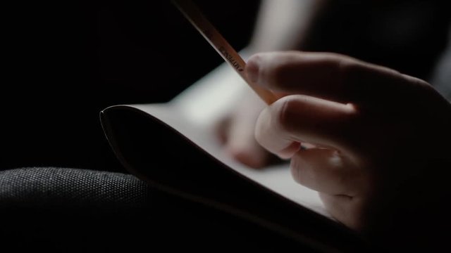 Close-up man's hand knocks nervously on the notebook with pencil and starts writing on dark background. Person thinks at the cafe. Guy is considering a solution to the problem