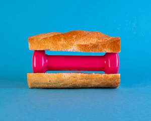 Sandwich with pink fitness dumbbell side view. Hand weight between bread slices on blue background. Eating baguette and training. Balanced nutrition, dieting concept. Carbohydrate, carbs consumption