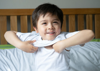 School boy sitting in bed and try to wearing his cloth with smiling face,Cute kid boy getting...