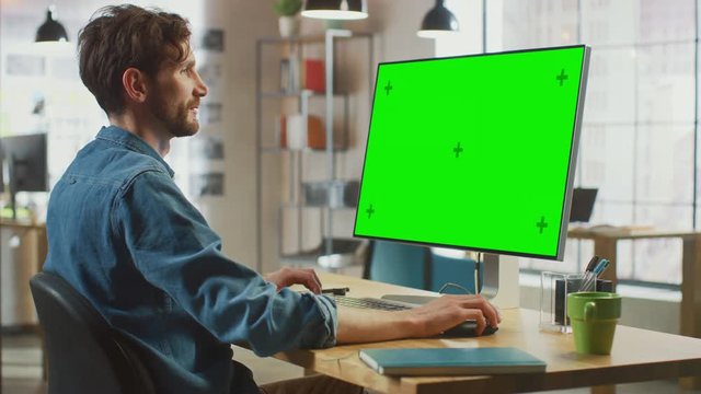 Male Creative Designer with Beard and Jeans Shirt Works on His Personal Computer with Big Green Screen Mock Up Display. He Works in Cool Office Loft. Female Creative Colleague Walks in Background.