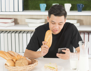 Young Asian man eating breakfast and playing smartphone in kitchen room, Freelance businessman