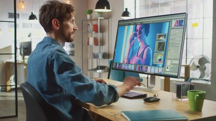 Male Digital Editor in Jeans Shirt Works in Photo Editing Software on His Personal Computer with Big Display. He Works in Cool Office Loft. Other Female Creative Colleague Walks in the Background.