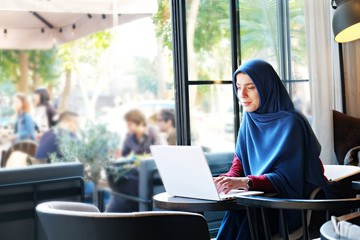 Young beautiful caucasian woman wearing traditional muslim headscarf in hipster coffee shop with big full length windows. Female in blue hijab at cozy cafe. Background, copy space, close up portrait.