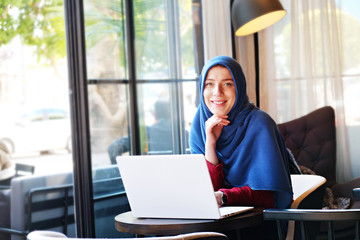 Fototapeta na wymiar Young beautiful caucasian woman wearing traditional muslim headscarf in hipster coffee shop with big full length windows. Female in blue hijab at cozy cafe. Background, copy space, close up portrait.
