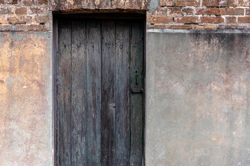 Close up of an old textured and weathered dark grey and brown wooden entrance door with stained concrete and brick wall