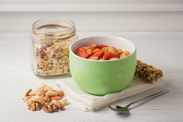 Granola cereal flakes with dried fruit, nuts in green bowl on white wooden table.