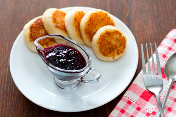 Cheese pancakes with currant jam, on a white plate with a fork, a teaspoon and a napkin, view from the top.