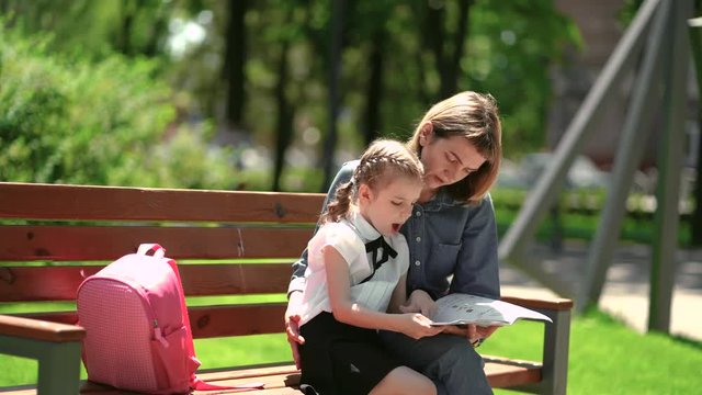 Mother and little daughter in outdoors public park sitting on the bench and read book, study lessons. Parenthood and child concept. Mother kissing her child.