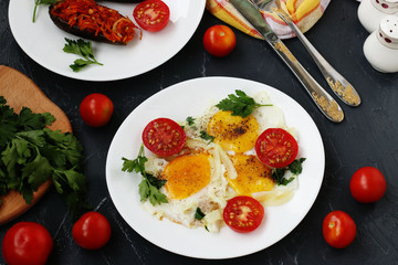 Scrambled eggs with cherry tomatoes are located on a white plate on a dark background, the photo there baked eggplants, parsley, cutlery, top view
