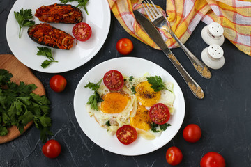 Scrambled eggs with cherry tomatoes are located on a white plate on a dark background, the photo there baked eggplants, parsley, cutlery, top view