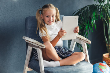Technologies, people concept - young blondy girl sitting on a chair and watching the tablet or surfing the net and smiling.