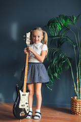 young blonde girl with tails in white t-shirt, skirt and sandals with electric guitar at home
