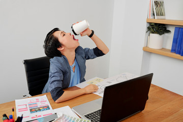 Asian young office worker sitting at her workplace with documents and laptop computer and drinking coffee from disposable cup to the bottom