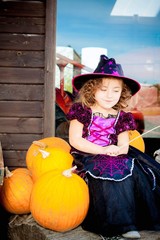 Happy Halloween! Cute cheerful little witch with a magic wand. Beautiful child girl in witch costume sitting on the big pumpkin, conjuring and laughing.