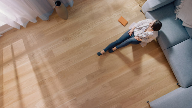 Young Woman is Sitting on a Floor and Looking out of the Window after Reading a Book. Cozy Living Room with Modern Interior, Grey Sofa and Wooden Flooring. Top View Camera Shot.