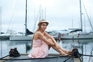 brown-haired smiling woman in a hat sits on the pier, looks at the beautiful yachts, travel concept