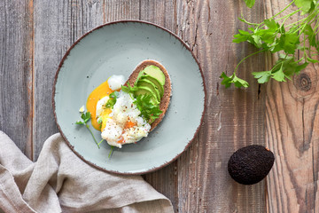 Avocado sandwich with dark bread, sliced avocado and poached egg, flat lay on rustic wood