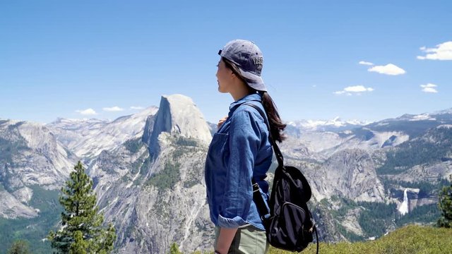 Young smiling chinese woman hiker manage to gets to top standing in front of beautiful dolomites and blue sky landscape in yosemite surroundings. female backpacker enjoy half dome trail view outdoor