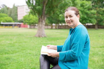 Obraz premium Happy relaxed woman with book enjoying weekend outdoors. Serene Caucasian lady sitting in park, reading novel and smiling at camera. Hobby concept
