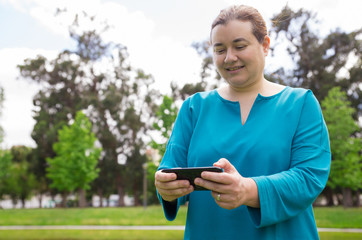 Fototapeta premium Smiling pensive woman watching media content on phone outdoors. Middle aged Caucasian lady using smartphone while walking in park. Wireless connection concept