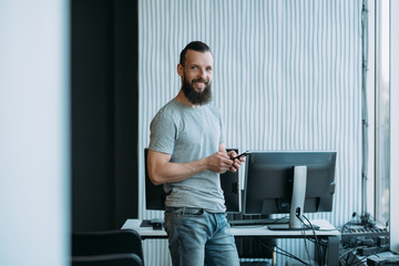Computer programming. Portrait of successful software developer standing at office, holding phone, smiling. Copy space.