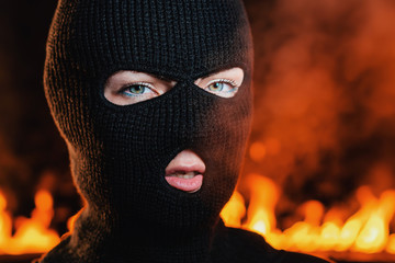 Portrait of young woman in black balaclava against backdrop of a blazing night fire. Concept of mass rallies and riots
