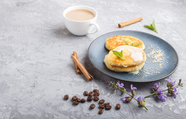 Cheese pancakes on a blue ceramic plate and a cup of coffee on a gray concrete background.