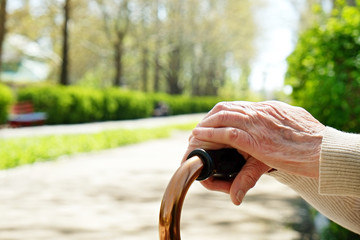 Senior disabled caucasian woman hands on cane outside nursing home park. Close up of elderly lady holding a walking stick outdoors of healthcare facility on the sunny day.