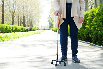 Senior disabled caucasian woman hands on cane outside nursing home park. Close up of elderly lady holding a walking stick outdoors of healthcare facility on the sunny day.