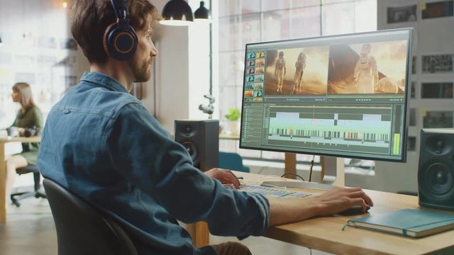 Male Video Editor Puts Headphones and Starts Working with Footage on His Personal Computer with Big Display. He Works in a Cool Office Loft. Creative Man Wears a Jeans Shirt.