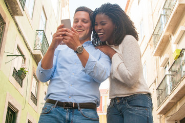 Positive multi-ethnic couple laughing while using mobile app on smartphone. Jolly excited young tourists enjoying vacation in city. Technology concept