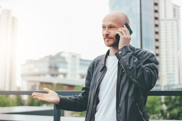 Adult attractive bald bearded man talking by mobile at city street