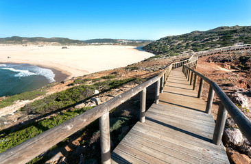 Fototapeta na wymiar Path on hills over ocean, made from wood, and empty windy sunny beach in Portugal town. Ocean waters and green hills over peaceful seaside at sunny day