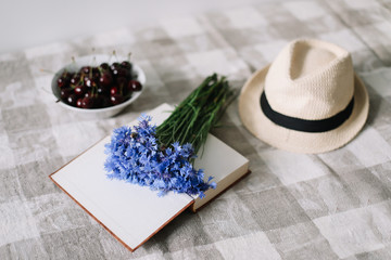 Obraz na płótnie Canvas Fresh cherries, cornflowers, straw hat and a book on the table. Summer flat lay wallpaper, top view 