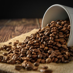 Coffee beans overflowing from a white coffee cup placed on a sackcloth and a black background.